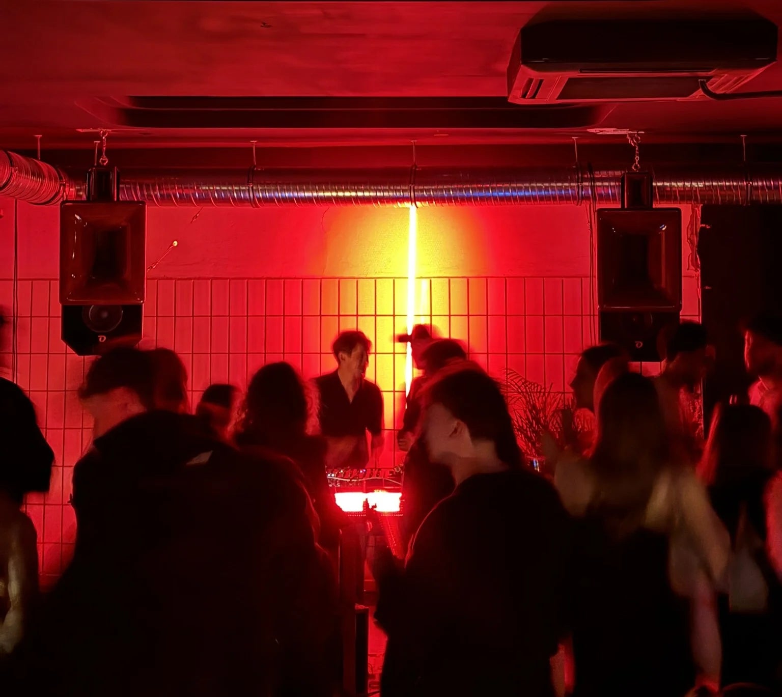 People in a club setting with red lighting and a DJ booth.