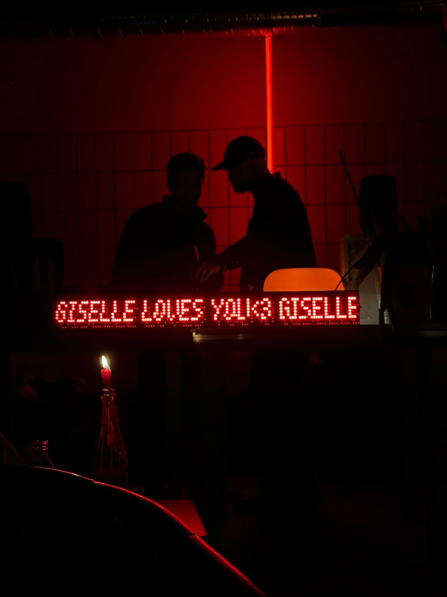 Two people sitting at a table with a red neon sign in front of them displaying 'GISELLE LOVES YOU & RISELLE'.