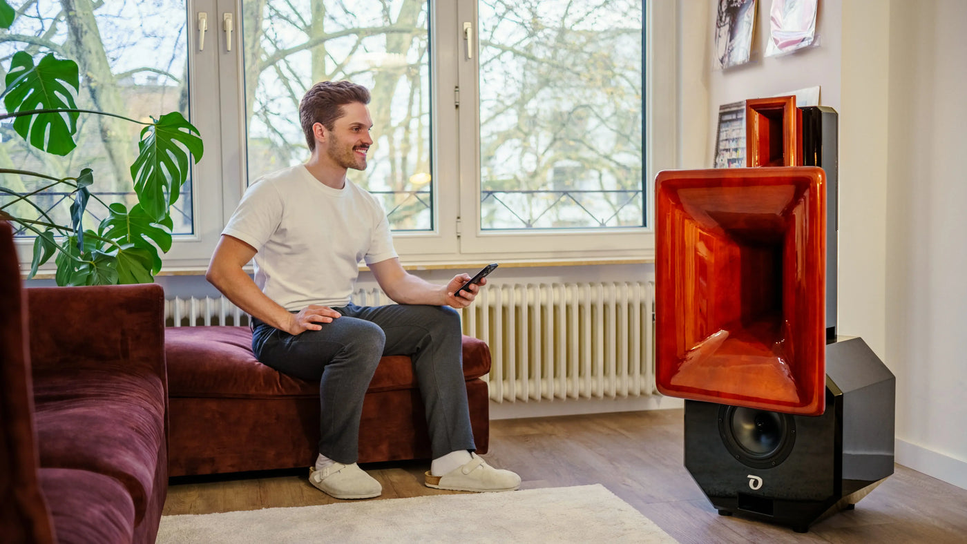 Man sitting on a couch using a smartphone next to a large red speaker in a room with windows and plants.